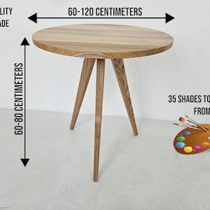 May include: A round wooden table with three legs. The table is 60-120 centimeters wide and 60-80 centimeters tall. The table is made of natural wood and has a light brown color. The text "HIGH QUALITY HANDMADE" is visible in the top left corner of the image. The text "60-120 CENTIMETERS" is visible above the table. The text "60-80 CENTIMETERS" is visible to the left of the table. The text "35 SHADES TO CHOOSE FROM" is visible in the bottom right corner of the image.