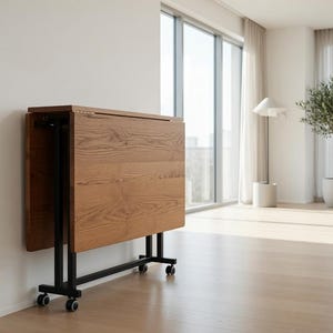 May include: A foldable table with a rectangular wooden top featuring a visible grain pattern. The table is supported by a black metal frame with four small wheels at the base, allowing it to be folded vertically for compact storage against a wall.