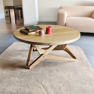 Op de afbeelding: Een ronde houten salontafel met een natuurlijke houten afwerking en een unieke X-vormige basis. De tafel is versierd met een stapel boeken en een rode kaars.