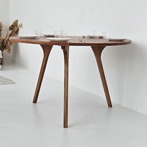 Pode incluir: Uma mesa de jantar redonda de madeira com um design &uacute;nico de tr&ecirc;s pernas. A mesa &eacute; feita de madeira castanha escura e est&aacute; montada com pratos brancos e copos transparentes. As pernas da mesa t&ecirc;m uma forma curva distinta. A mesa tem cerca de 122 cm de di&acirc;metro.