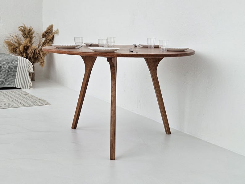 Pode incluir: Uma mesa de jantar redonda de madeira com um design &uacute;nico de tr&ecirc;s pernas. A mesa &eacute; feita de madeira castanha escura e est&aacute; montada com pratos brancos e copos transparentes. As pernas da mesa t&ecirc;m uma forma curva distinta. A mesa tem cerca de 122 cm de di&acirc;metro.
