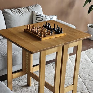May include: A wooden folding table with a chessboard set on top. The table is light brown and has a simple design. The chessboard is black and white with black and white chess pieces.