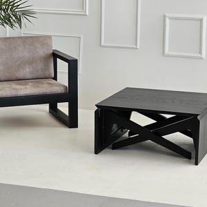 May include: A black wooden coffee table with a unique folding design. The table has a rectangular top and a black frame with a geometric pattern. The table is sitting on a white floor.
