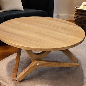 May include: A round wooden coffee table with a light brown finish and a unique X-shaped base. The table is sitting on a light brown rug.