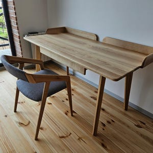 Könnte beinhalten: Ein heller Holzschreibtisch mit einem grauen gepolsterten Stuhl. Der Schreibtisch hat ein schlichtes Design mit einer rechteckigen Tischplatte und zwei Beinen auf jeder Seite. Der Stuhl hat eine geschwungene Rückenlehne und einen bequemen Sitz.
