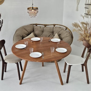 Pode incluir: Uma mesa de jantar redonda de madeira com quatro cadeiras, pronta para uma refei&ccedil;&atilde;o. A mesa tem um acabamento em madeira natural e est&aacute; rodeada por pratos brancos, copos e talheres. Um sof&aacute; de vime e elementos decorativos completam o ambiente.