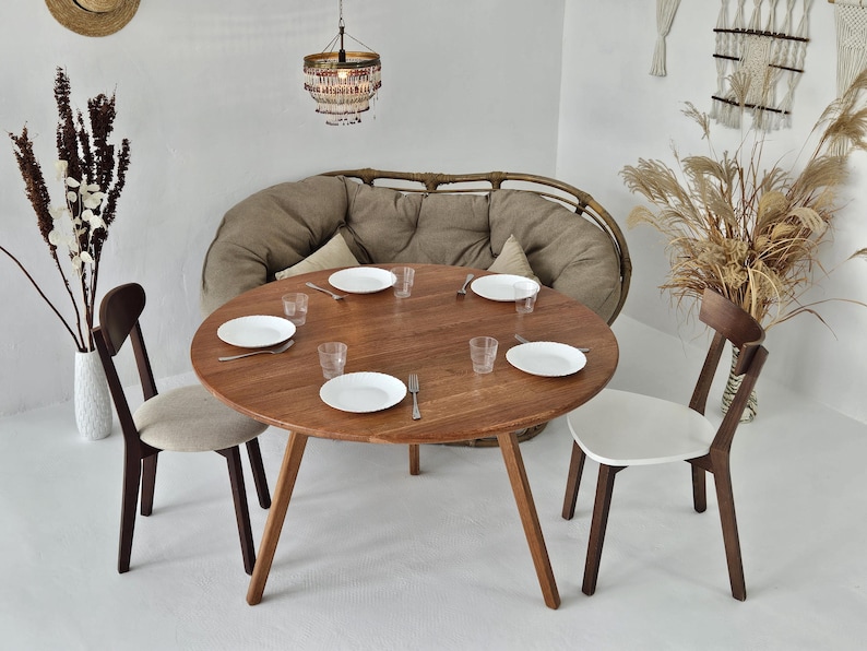 Pode incluir: Uma mesa de jantar redonda de madeira com quatro cadeiras, pronta para uma refei&ccedil;&atilde;o. A mesa tem um acabamento em madeira natural e est&aacute; rodeada por pratos brancos, copos e talheres. Um sof&aacute; de vime e elementos decorativos completam o ambiente.