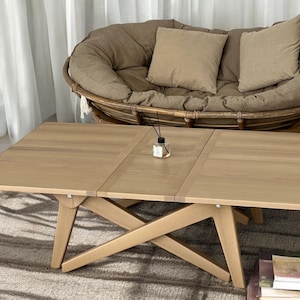 Può includere: Un tavolino da caffè in legno chiaro con una base a forma di croce e un piano rettangolare. Il tavolo è composto da quattro pezzi separati che possono essere ripiegati insieme. Il tavolo è posizionato su un tappeto marrone chiaro davanti a una sedia in vimini con cuscini.