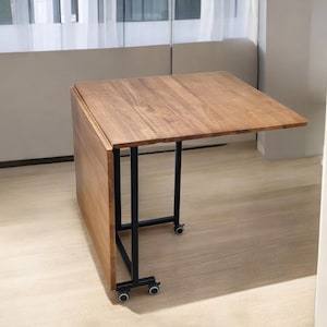 Peut inclure: Table pliante en bois avec un plateau rectangulaire et des pieds en métal noir. La table est partiellement pliée, révélant sa conception à charnière. La table est sur roulettes pour faciliter le déplacement.