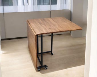 May include: A wooden folding table with a rectangular top and black metal legs. The table is partially folded, revealing its hinged design. The table is on wheels for easy movement. The wood has a warm, natural tone.