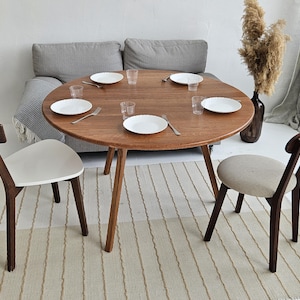 Pode incluir: Uma mesa de jantar redonda de madeira com tr&ecirc;s pernas, com pratos brancos, copos e talheres. Duas cadeiras com estrutura de madeira e assentos brancos e bege est&atilde;o posicionadas ao redor da mesa. Um tapete de tons neutros est&aacute; por baixo.