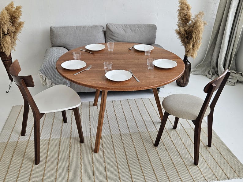 Pode incluir: Uma mesa de jantar redonda de madeira com tr&ecirc;s pernas, com pratos brancos, copos e talheres. Duas cadeiras com estrutura de madeira e assentos brancos e bege est&atilde;o posicionadas ao redor da mesa. Um tapete de tons neutros est&aacute; por baixo.
