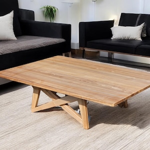 May include: A light brown wooden coffee table with a rectangular top and a unique X-shaped base. The table is sitting on a light beige rug in front of a black sofa.