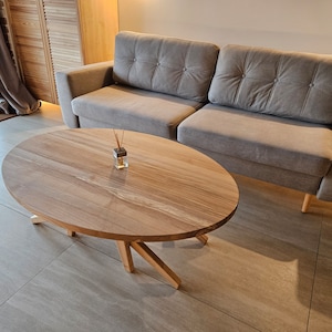 May include: A light brown oval wooden coffee table with a natural wood grain finish and a simple, modern design. The table has four legs that are angled outwards and a smooth, flat top surface. The table is sitting on a light beige tiled floor.