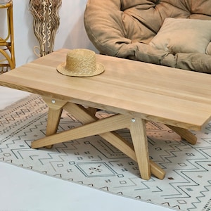 May include: A light wood coffee table with a unique X-shaped base. The table has a natural wood finish and is sitting on a patterned rug.