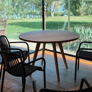 May include: A round wooden table with four black chairs sits in a room with large windows overlooking a grassy area. The table has a natural wood finish and the chairs have a woven design.