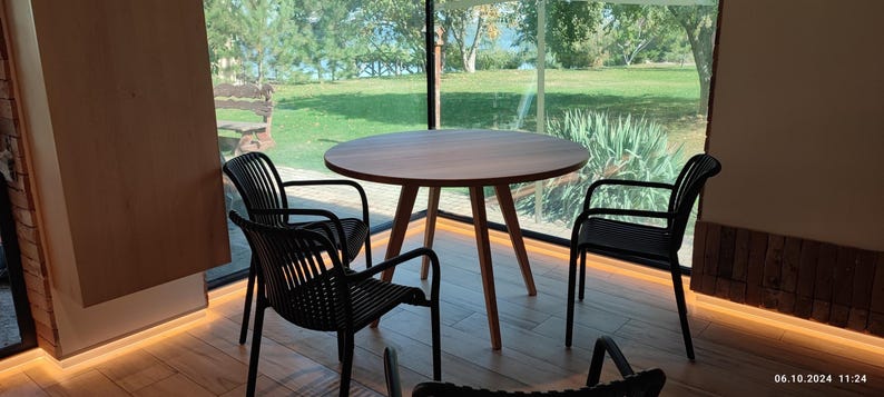 May include: A round wooden table with four black chairs sits in a room with large windows overlooking a grassy area. The table has a natural wood finish and the chairs have a woven design.