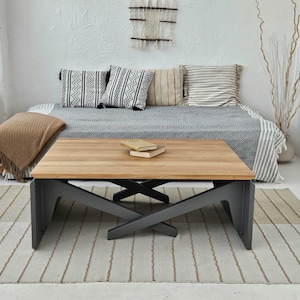 Könnte beinhalten: Ein Holz-Couchtisch mit schwarzem Metallgestell. Der Tisch hat eine helle Holzplatte und ein Kreuzdesign an den Beinen. Der Tisch steht auf einem grau-weiß gestreiften Teppich.