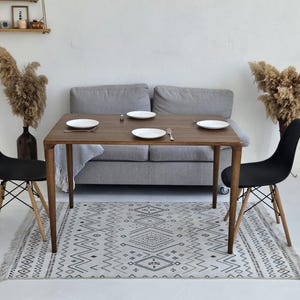 Könnte beinhalten: Esszimmer-Szene mit einem rechteckigen Holztisch, vier weißen Tellern, Besteck und einem Salzstreuer. Zwei schwarze Stühle mit Holzbeinen und ein gemusterter Teppich vor einem grauen Sofa. Dekorative Trockenpflanzen in Vasen.
