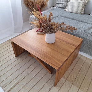 May include: A rectangular wooden coffee table with a natural finish. The table has a unique leg design and is placed on a striped rug. A white vase with dried flowers sits on top of the table. The table measures approximately 36 inches long.