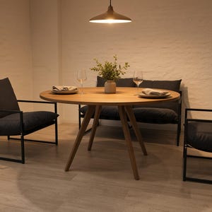 Könnte beinhalten: Ein runder Esstisch aus Holz mit vier schrägen Beinen, umgeben von zwei Sesseln mit schwarzem Metallrahmen und einer passenden schwarzen Bank. Der Tisch ist mit zwei Tellern, Servietten und Weingläsern sowie einer kleinen Vase mit Grün gedeckt.