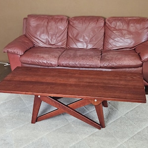 May include: A dark brown wooden coffee table with a unique X-shaped base. The table is in front of a brown leather sofa.