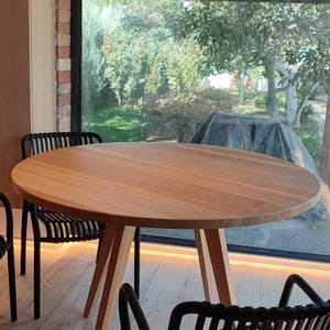 May include: A round wooden table with a natural wood finish and three legs. Two black chairs with woven seats are positioned around the table. The table is in front of a large window with a view of a green lawn and trees.