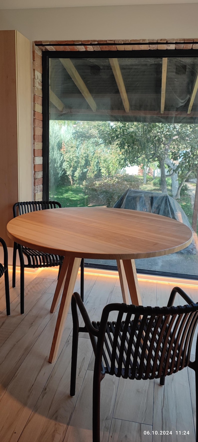 May include: A round wooden table with a natural wood finish and three legs. Two black chairs with woven seats are positioned around the table. The table is in front of a large window with a view of a green lawn and trees.