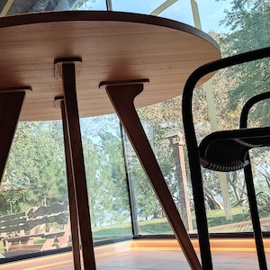 May include: A round wooden table with four legs sits in a room with large windows. Two black chairs with metal frames are positioned around the table.
