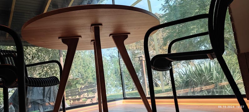 May include: A round wooden table with four legs sits in a room with large windows. Two black chairs with metal frames are positioned around the table.