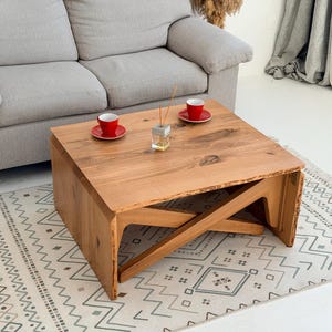 May include: A square wooden coffee table with a natural wood finish. The table has a unique cross-beam design on the sides. Two red cups and saucers sit on top, along with a small glass diffuser. A gray sofa and patterned rug are in the background.