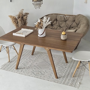 May include: A wooden dining table with a light brown finish and a white patterned rug underneath. Two white chairs are placed on either side of the table. A small white planter with a plant is on the table, along with a decorative tray and a small gold candle holder.