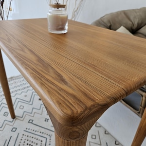 Puede incluir: Una mesa de madera marrón claro con bordes y patas redondeadas. La mesa tiene un patrón de vetas de madera natural y un pequeño frasco de vidrio con una vela en su interior. La mesa se utiliza probablemente para la decoración del hogar o como mesa auxiliar.