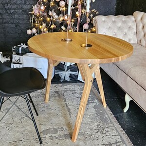 May include: A round wooden table with a light wood finish and three legs. The table is set with two candle holders and is surrounded by a black chair and a light beige tufted sofa.