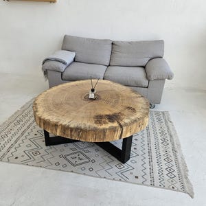 Op de afbeelding: Een ronde houten salontafel met een natuurlijke houtnerf afwerking, ondersteund door een zwart metalen onderstel. De tafel staat op een patroonkleed. Een grijze bank is op de achtergrond te zien.