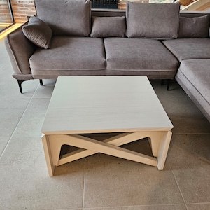 May include: A white coffee table with a unique X-shaped base. The table is made of wood and has a light wood finish. The table is sitting in front of a gray sectional sofa.