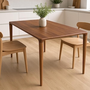 May include: A rectangular wooden dining table with a dark brown finish and four tapered legs, accompanied by two light-coloured wooden chairs with curved backrests. A white ceramic vase containing a green plant sits on the table.