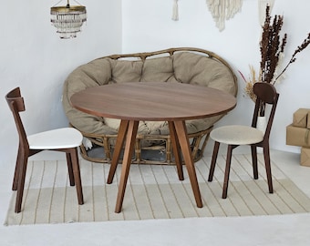 May include: A round wooden table with a brown top and three legs. Two chairs with white seats are placed around the table. The chairs are made of wood and have a brown finish.