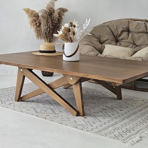 May include: A wooden coffee table with a rectangular top and a unique X-shaped base. The table is made of dark brown wood and has a natural finish. The table is sitting on a white rug with a geometric pattern.