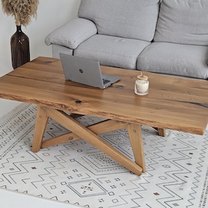 May include: A rectangular wooden coffee table with a natural wood finish. The table has a live edge and sits on a crossed-leg base. A laptop and a candle are on the table. The table is 152 cm long.