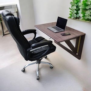 Op de afbeelding: Een donkerbruin houten wandbureau met een laptop en een telefoon op het oppervlak. Een zwarte bureaustoel met wielen staat voor het bureau.
