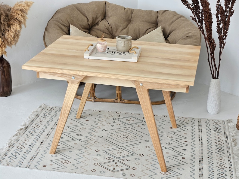 May include: A light wood table with a rectangular top and four legs. The table is sitting on a patterned rug in front of a brown chair.