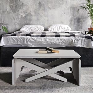 May include: A white wooden coffee table with an X-shaped base sits on a gray rug. Two books rest on the table's surface. A bed with white bedding and a black and white blanket is in the background.