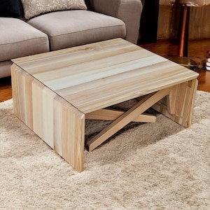 May include: A light-coloured wooden coffee table with a rectangular top and a unique, geometric base. The table is made of light-coloured wood and sits on a beige rug. The table's design features a series of vertical wooden slats.