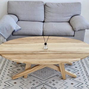 Könnte beinhalten: Ein ovaler Couchtisch aus hellem Holz mit natürlicher Oberfläche und einem einzigartigen X-förmigen Gestell. Der Tisch steht auf einem weißen und grauen gemusterten Teppich.