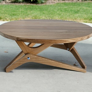 May include: A round wooden coffee table with a unique X-shaped base. The table is made of light brown wood and has a smooth, polished finish.