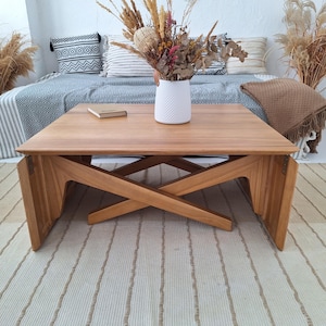 Op de afbeelding: Een lichtbruine houten salontafel met een X-vormige basis. De tafel heeft een rechthoekig blad en is omgeven door een vloerkleed, kussens en een vaas met gedroogde bloemen. De tafel is ongeveer 100 cm lang.