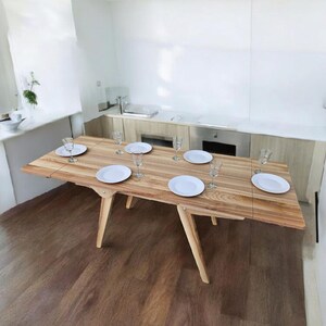 May include: A wooden dining table with a light wood finish and six white plates and glasses set for a meal. The table is set against a white wall and has a light wood floor.