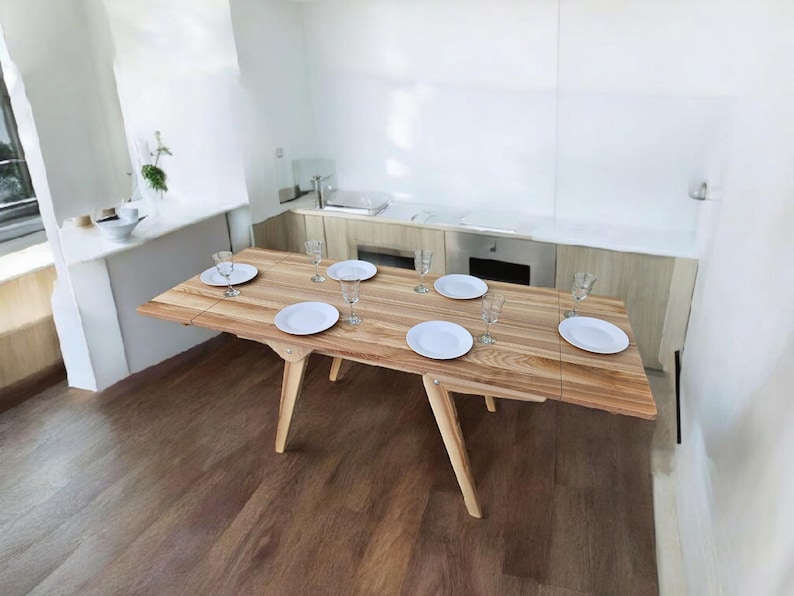 May include: A wooden dining table with a light wood finish and six white plates and glasses set for a meal. The table is set against a white wall and has a light wood floor.