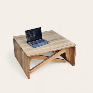 Op de afbeelding: Een lichtbruine houten salontafel met een laptop erop. De tafel heeft een vierkant blad en een uniek ontwerp met schuine poten. De laptop is open en toont een donker scherm.